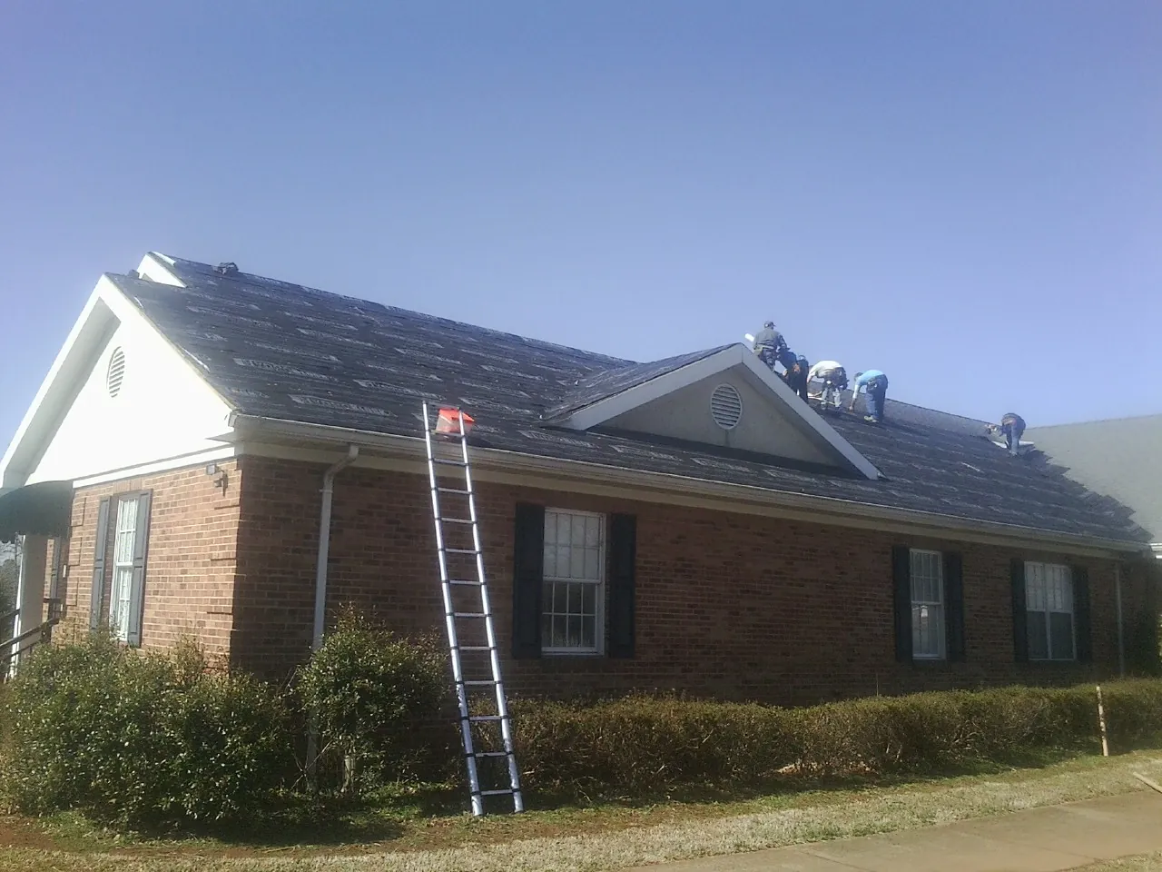 Crew tearing shingles off a brick ranch roof with a ladder set — Emergency Roof Repair work by our crew in Arcola