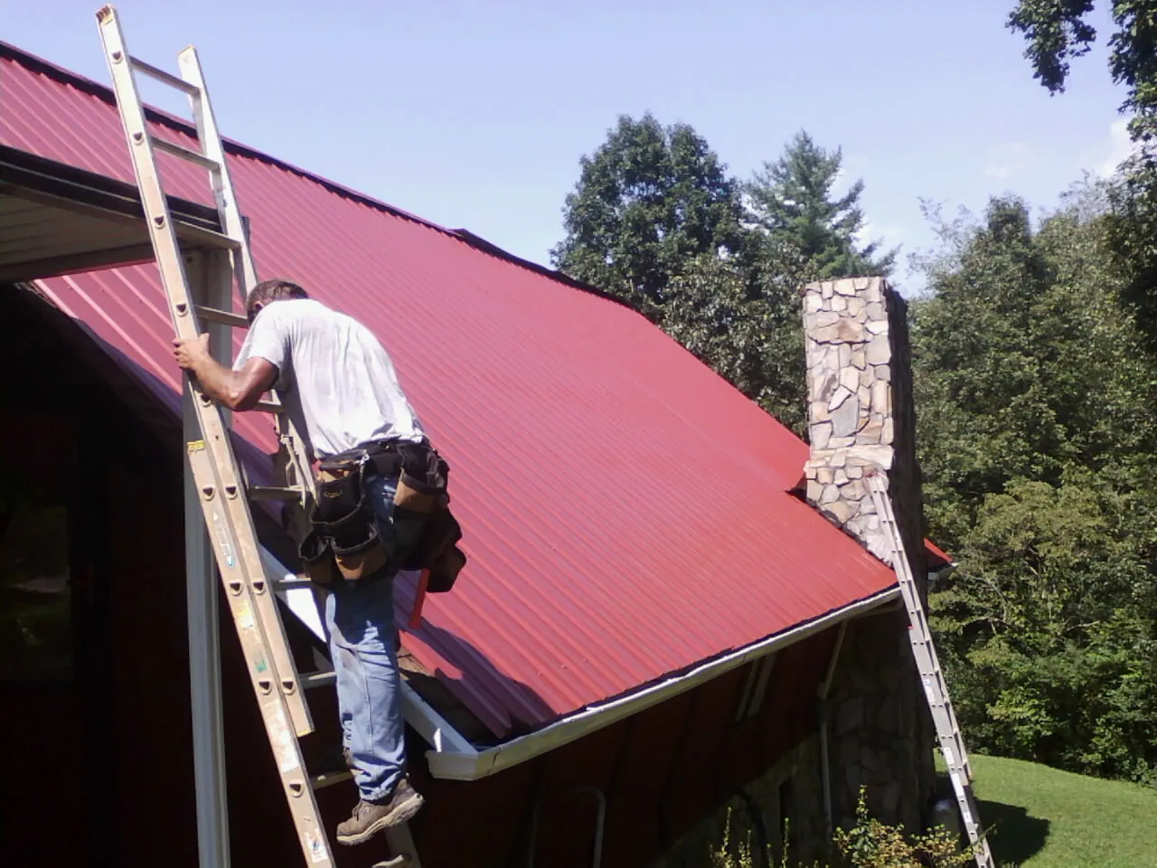 Roofer on a ladder beside a red metal roof and stone chimney — Soffit & Fascia Repair by Roofing Brothers in Arcola